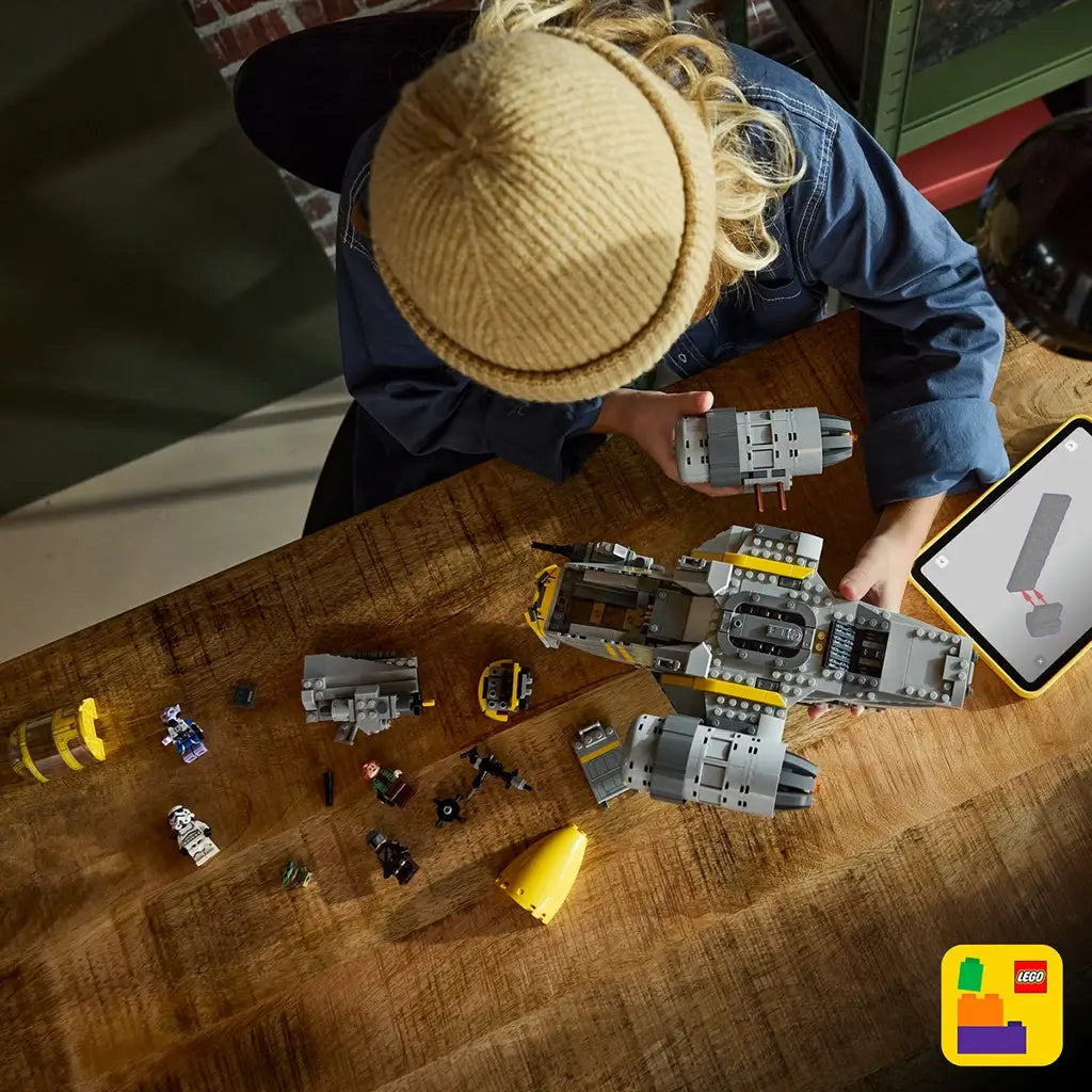 LEGO Star Wars Razor Crest construction set, gray and yellow plastic spacecraft with detailed engine modules and removable sections, shown during assembly on a wooden table beside a tablet guide.