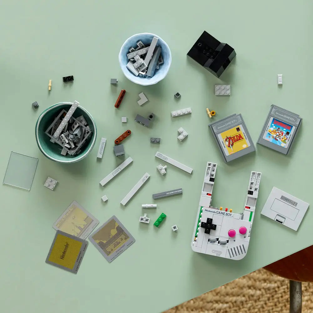 LEGO pieces for Nintendo Game Boy model spread across a light green table, showing gray bricks, cartridge elements, and interchangeable printed screen tiles mid-build.