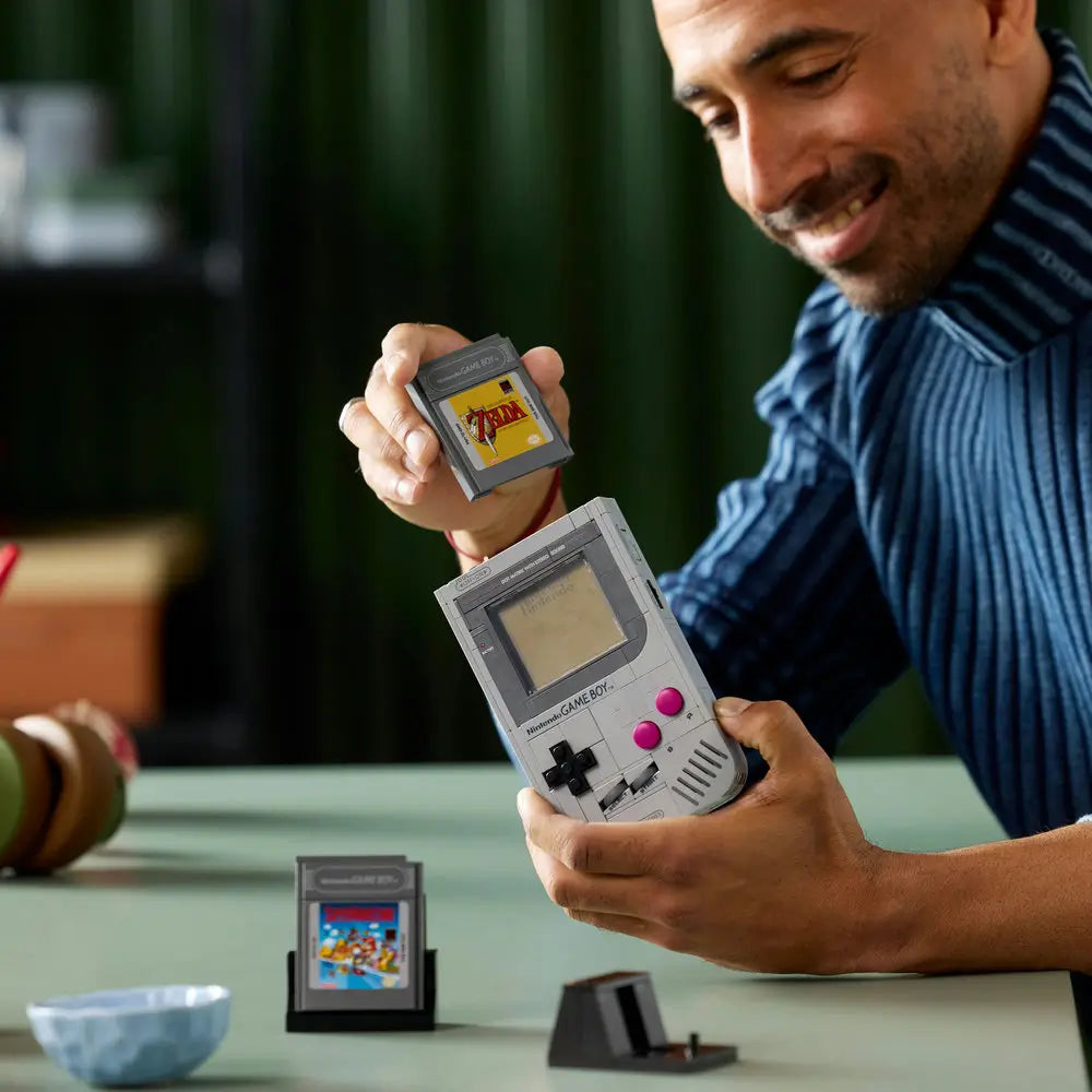 Smiling person assembling the LEGO Nintendo Game Boy model, inserting a brick-built The Legend of Zelda cartridge into the console replica during a hands-on build session.