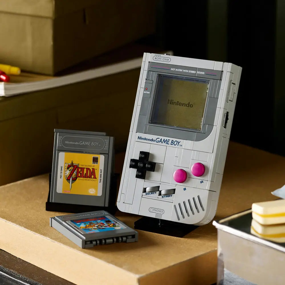 LEGO Nintendo Game Boy model displayed on a desk beside LEGO cartridges for The Legend of Zelda and Super Mario Land, built with gray, black, and pink LEGO bricks for nostalgic realism.
