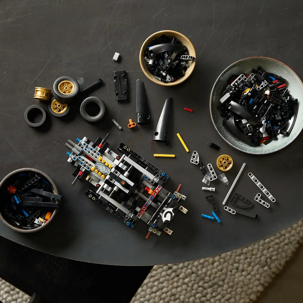 LEGO Technic Ford GT40 Mk II build activity shows black Technic beams, axles, gears, and gold wheel parts organized on a dark tabletop around a partially assembled chassis during focused construction.