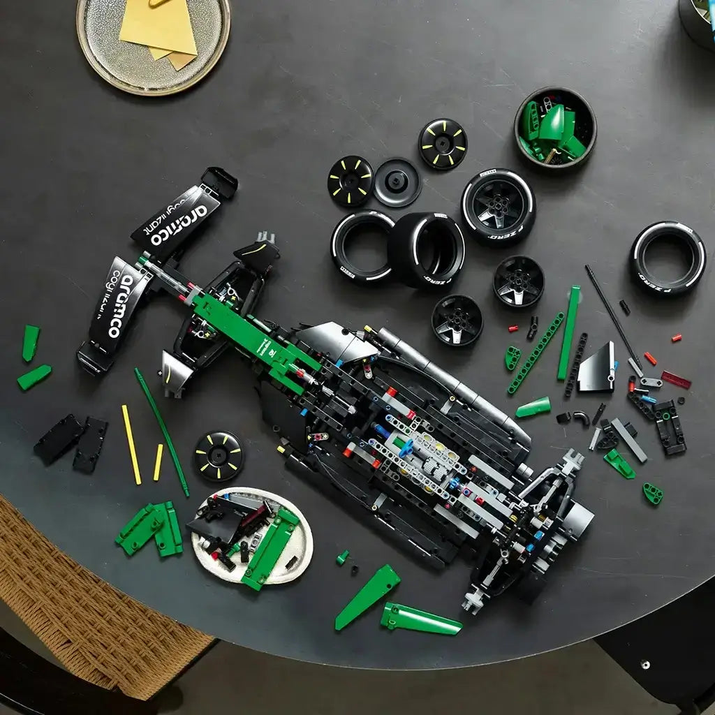 LEGO Technic Aston Martin Aramco AMR25 F1 car in green and black pieces is partly assembled on a dark table, surrounded by tires, body panels and mechanical components.