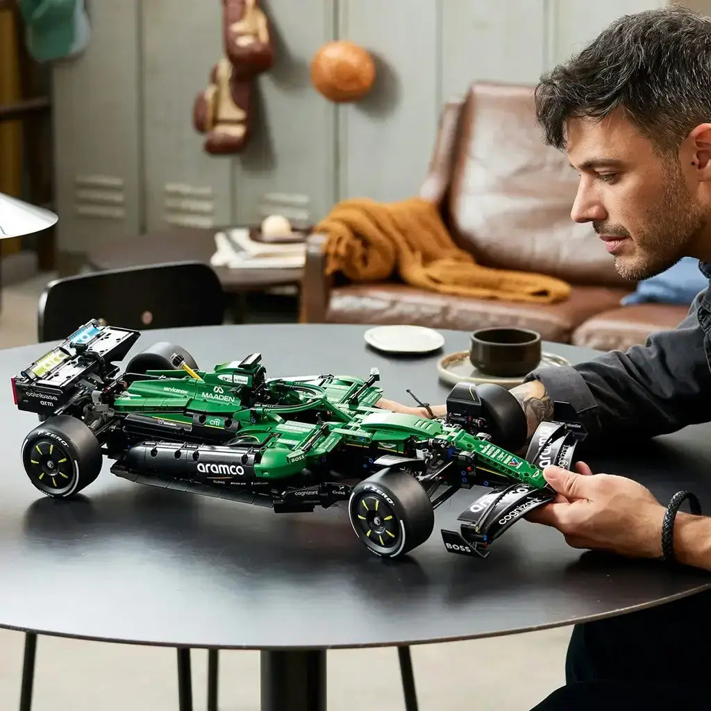 LEGO Technic Aston Martin Aramco AMR25 F1 car in green and black bricks sits on a dark table as an adult builder adjusts its detailed front wing in a living room.