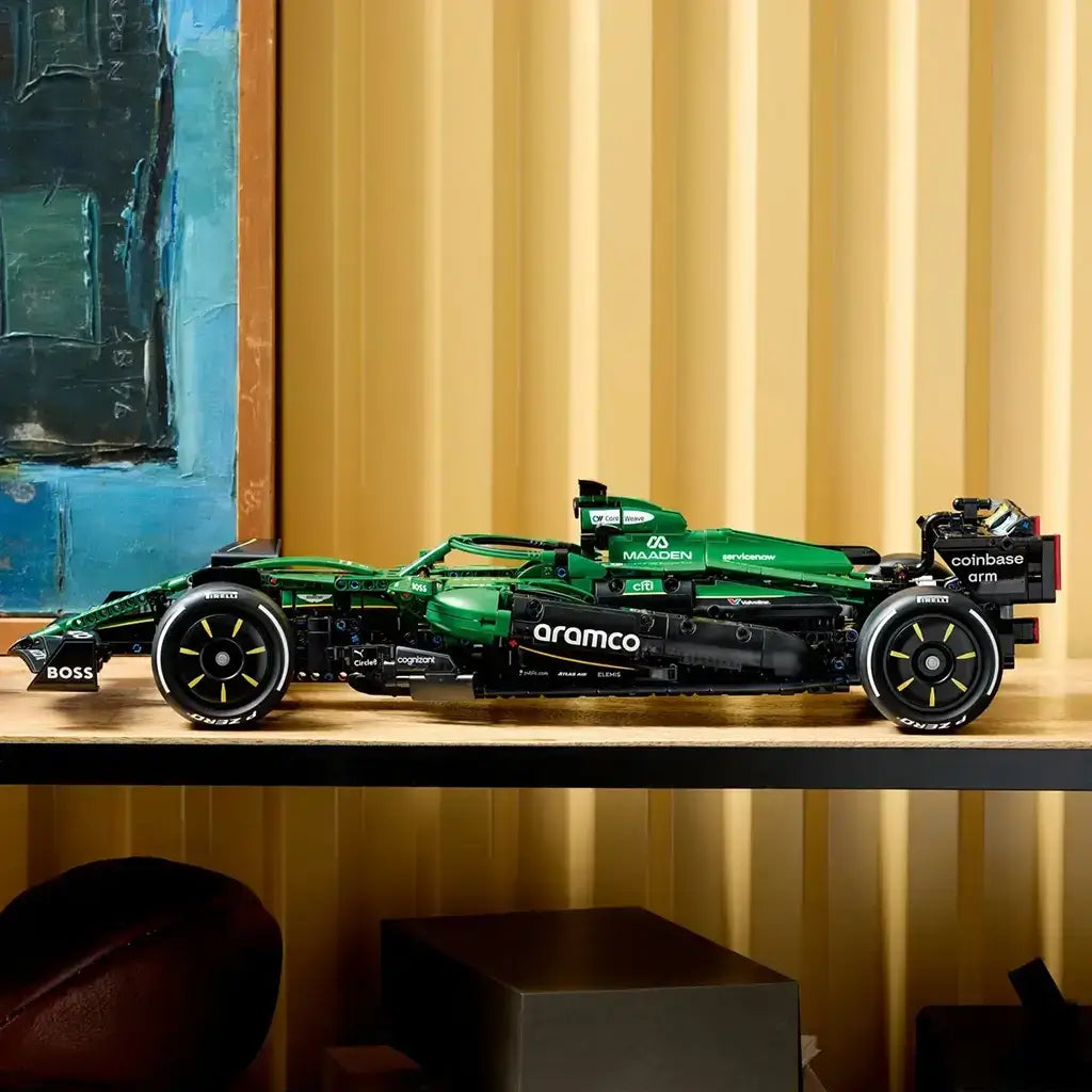 LEGO Technic Aston Martin Aramco AMR25 F1 car in green and black bricks is displayed in side profile on a shelf, with sponsor graphics and racing tires against a gold wall.