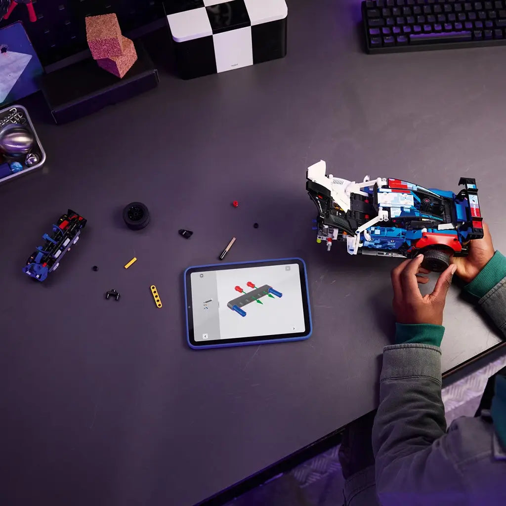 LEGO Technic BMW M4 GT3 EVO build scene shows multicolor plastic components and race car parts assembled on a desk, illustrating construction process with tools and digital instructions in a contemporary workspace setting.