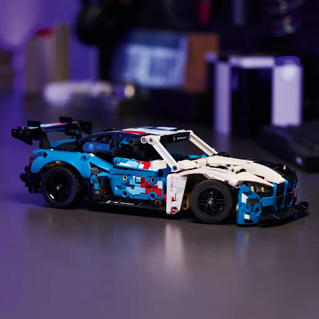 LEGO Technic BMW M4 GT3 EVO display shows a multicolor plastic race car with blue, white, black, and red panels, sleek low body, and rear wing, positioned on a tabletop in a softly lit indoor environment.