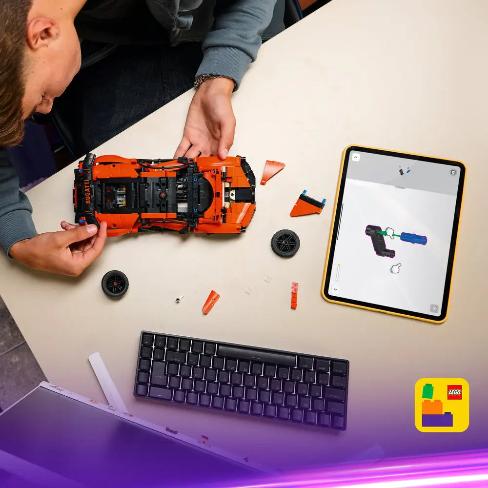 LEGO Technic Bugatti Chiron Pur Sport building scene shows orange Technic chassis and loose parts spread on a desk as a builder assembles the supercar using digital instructions on a tablet in a modern workspace.