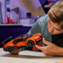 LEGO Technic Bugatti Chiron Pur Sport lifestyle image shows a builder closely inspecting the orange supercar model, adjusting front details and wheel alignment in a focused indoor building environment.