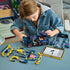 LEGO Technic Ford Mustang GT in dark blue plastic with pink and cyan details is customized by a young builder, with yellow body parts and a tablet on a worktable.