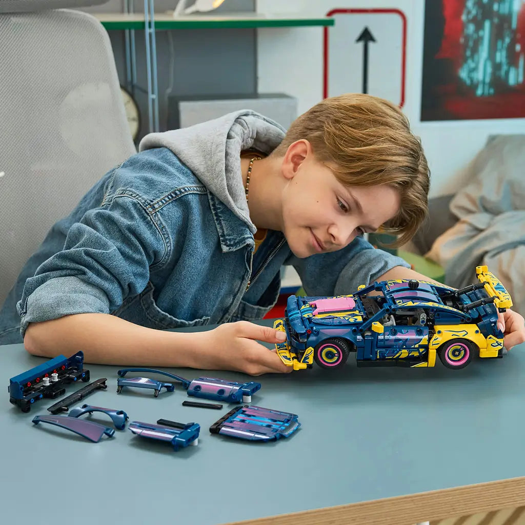 LEGO Technic Ford Mustang GT in dark blue plastic with pink and yellow custom panels is adjusted by a young builder, with alternate body parts spread across a worktable.