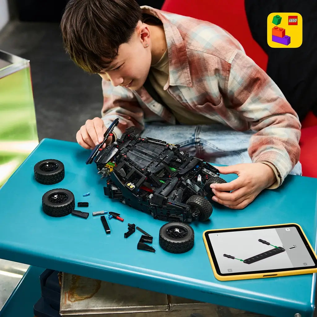 LEGO Technic Batmobile Tumbler build in black plastic sits partly assembled on a blue table as a young person works beside loose tires, pieces, and a tablet.