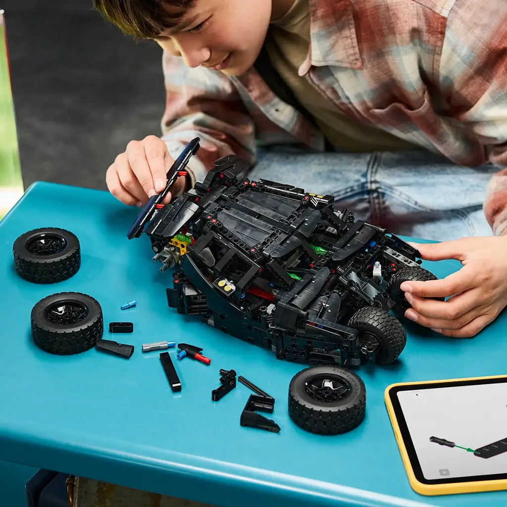 LEGO Technic Batmobile Tumbler build in black plastic is assembled by a young person at a blue table, with loose wheels, parts, and digital instructions on a tablet.