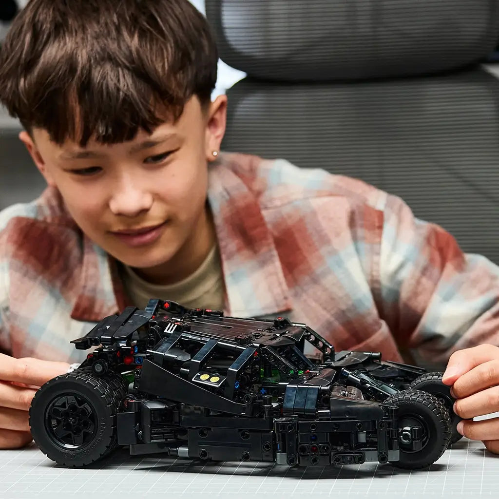 LEGO Technic Batmobile Tumbler model in black plastic is held by a young builder at a worktable, showing its low armored profile, rugged tires, and mechanical details.