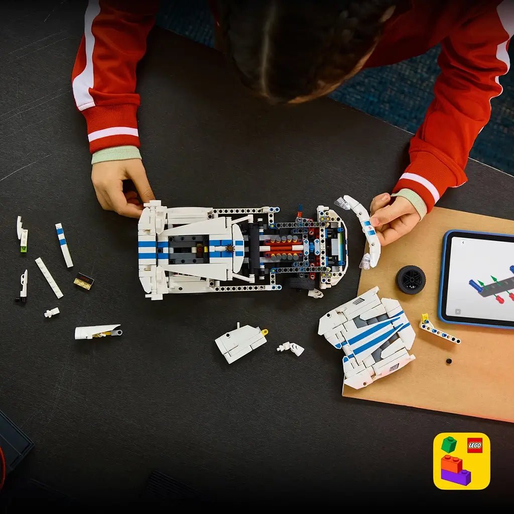 LEGO Technic Dodge Viper GTS-R in white plastic with blue stripes is assembled by a young builder, with body panels, loose parts and a tablet spread across a dark table.