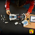 LEGO Technic Dodge Viper GTS-R in white plastic with blue stripes is assembled by a young builder, with body panels, loose parts and a tablet spread across a dark table.