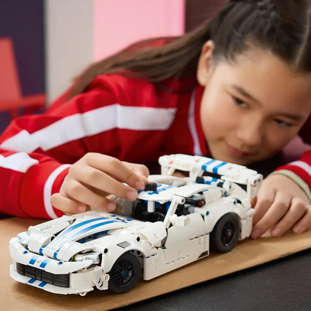 LEGO Technic Dodge Viper GTS-R in white plastic with blue racing stripes is adjusted by a young builder, showing black wheels and a large rear wing on a tabletop.