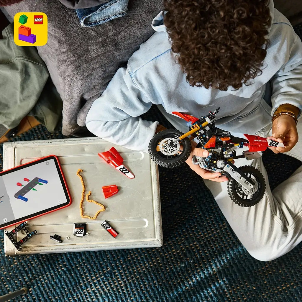 LEGO Technic Ducati Desmo450 MX Factory motorcycle in red and black is assembled by a young person beside loose parts and digital instructions on a tablet at home.
