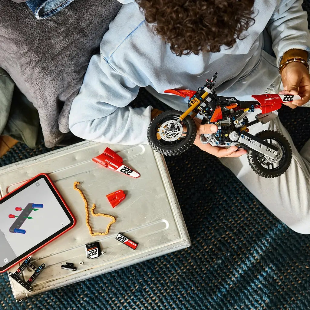 LEGO Technic Ducati Desmo450 MX Factory motorcycle in red and black is held by a young builder above a table with loose panels, chain pieces, and tablet instructions.