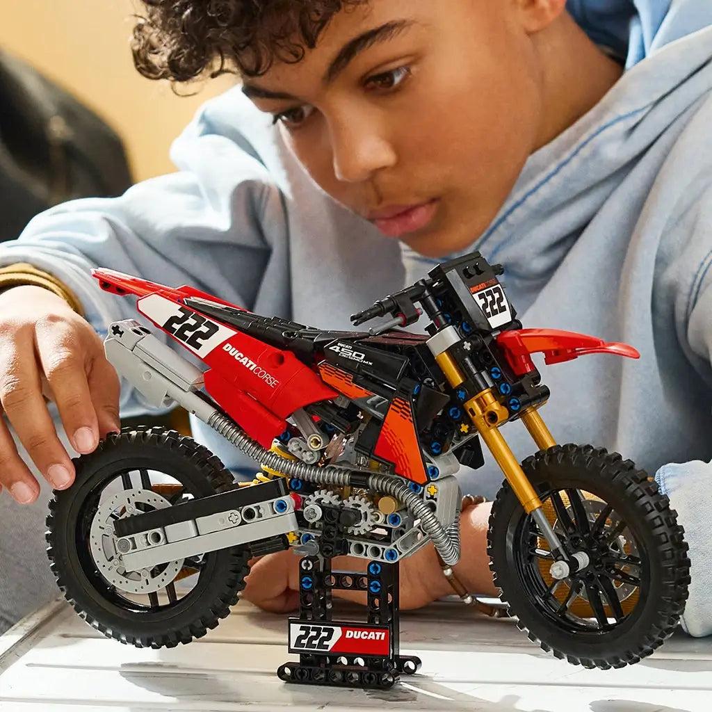 LEGO Technic Ducati Desmo450 MX Factory model in red, black, and gold stands on a display base as a young builder adjusts the rear wheel at a tabletop workspace.