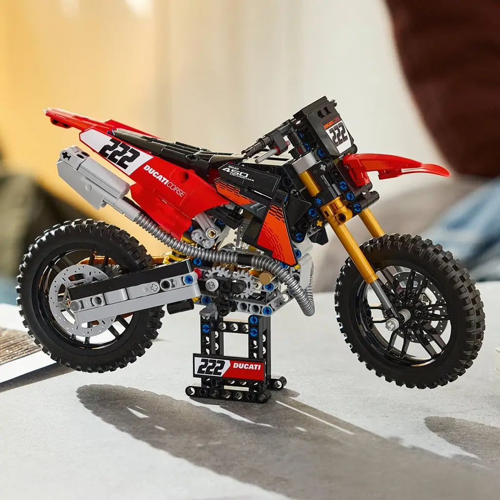 LEGO Technic Ducati Desmo450 MX Factory motorcycle in red, black, and gold features knobby tires and exposed gears on a display stand in a softly lit indoor setting.