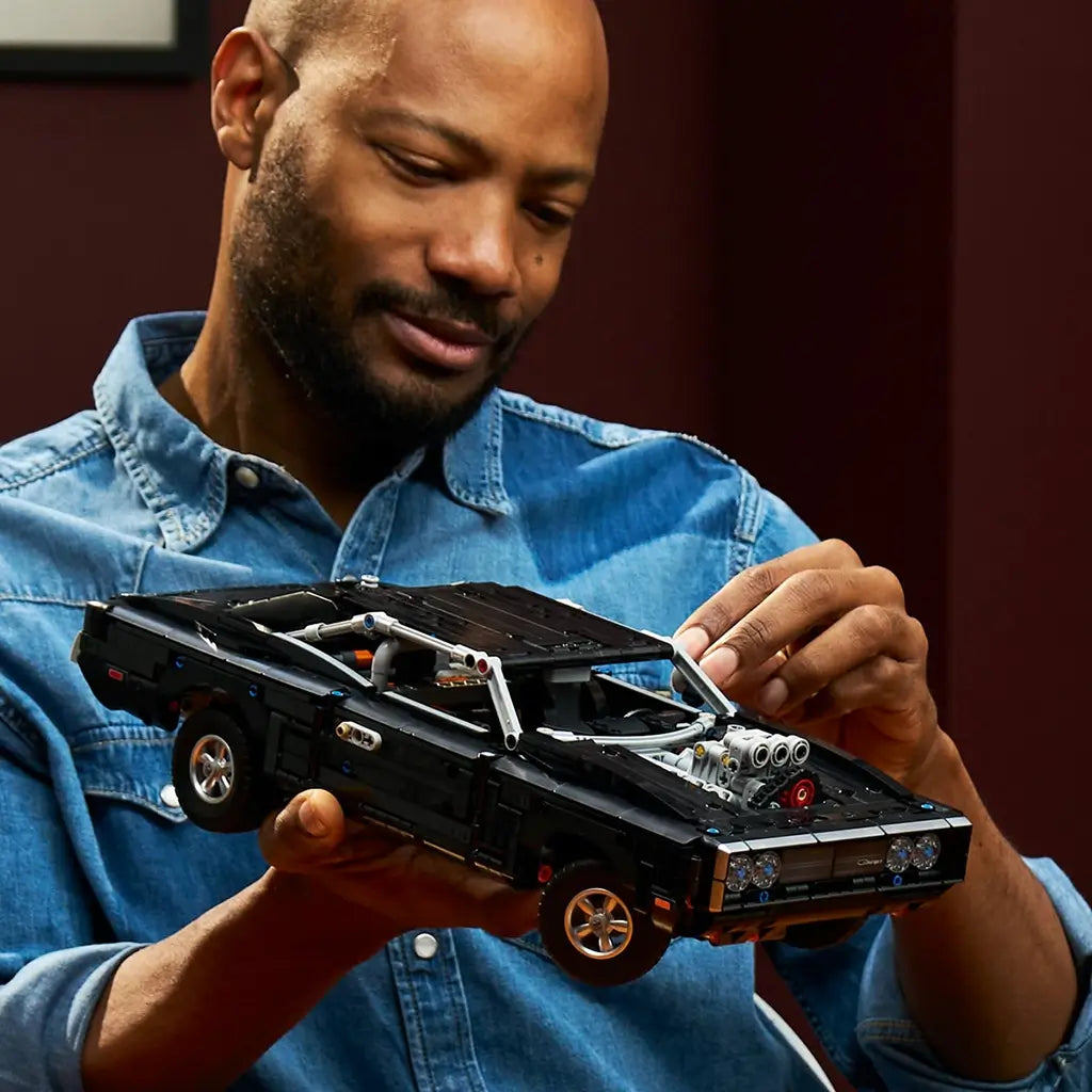 LEGO Technic Dodge Charger R/T in black plastic bricks with silver wheels and supercharger, held by an adult builder in a blue denim shirt against a dark red wall.