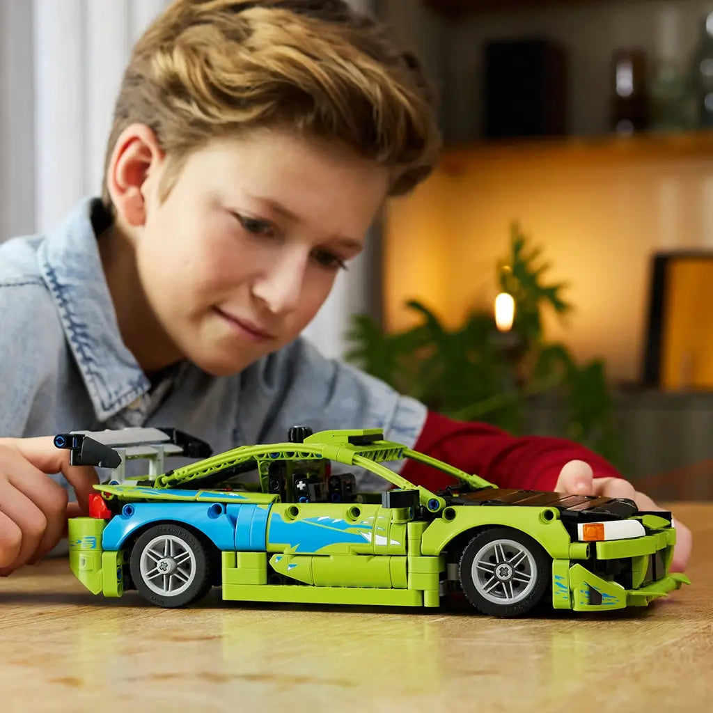 LEGO Technic Mitsubishi Eclipse in lime green and blue plastic bricks with silver wheels and rear wing, examined by a young builder at a wooden table in a warm room.