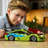 LEGO Technic Mitsubishi Eclipse in lime green and blue plastic bricks with silver wheels and rear wing, examined by a young builder at a wooden table in a warm room.