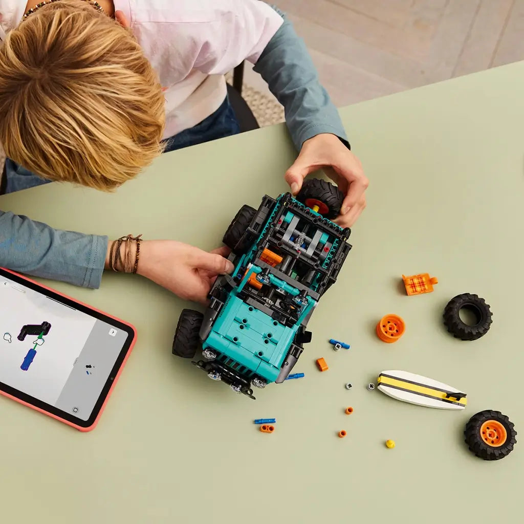 LEGO Technic Jeep Wrangler Rubicon build scene shows teal plastic vehicle components, black frame pieces, and orange details assembled on a table, illustrating construction process with tools and instructions in a casual indoor workspace.