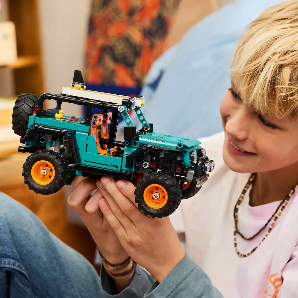 LEGO Technic Jeep Wrangler Rubicon toy shows a teal and black plastic SUV with textured tires and orange rims, held by a child while exploring features, representing hands on building play, set in a cozy indoor living room environment.