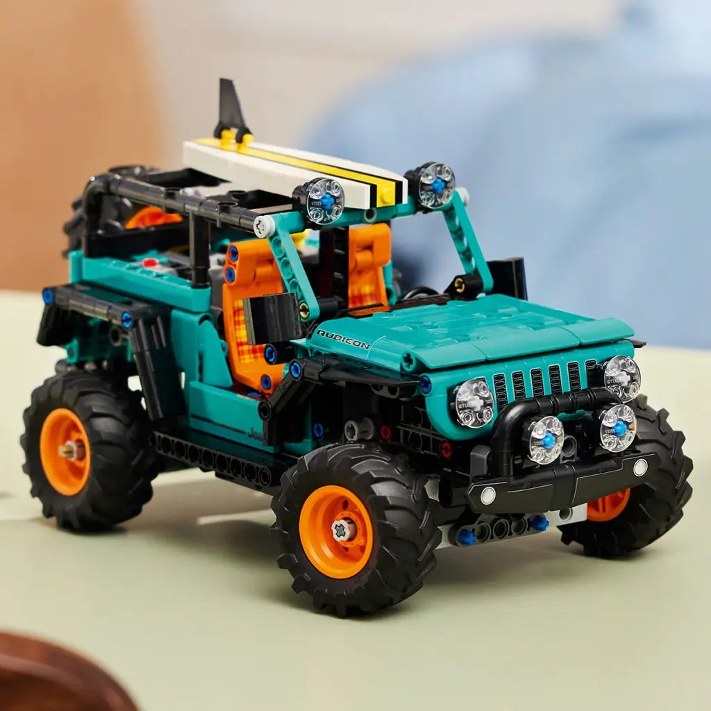 LEGO Technic Jeep Wrangler Rubicon display features a teal and black plastic SUV with rugged tires, orange rims, and detailed front lights, representing an off road vehicle model, placed on a tabletop in a softly lit home setting.
