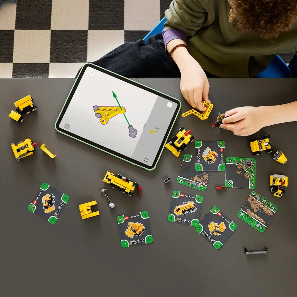 LEGO Technic Mighty Machines in yellow plastic surround a tablet and illustrated game cards as a young builder assembles a small construction vehicle at a gray table.
