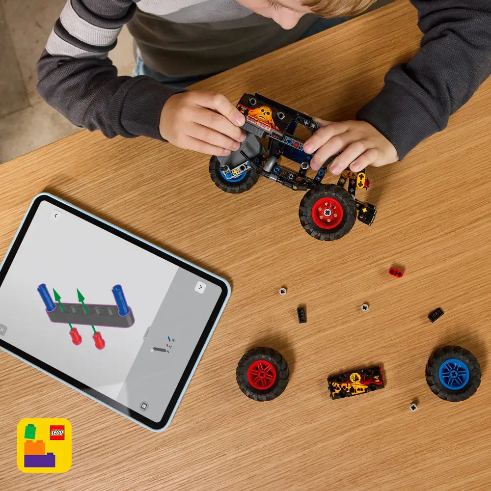 LEGO Technic Monster Jam Grave Digger build scene shows black Technic chassis, red and blue wheel parts, axles, and panels spread on a wooden table as a child assembles the pull-back truck using tablet instructions.