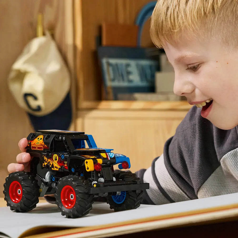 LEGO Technic Monster Jam Grave Digger lifestyle image shows a smiling child pushing the finished monster truck across a tabletop, emphasizing oversized tires, bold graphics, and hands-on pull-back play.