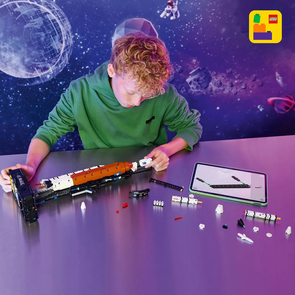 LEGO Technic NASA Artemis Space Launch System build scene shows white and orange Technic rocket sections laid horizontally on a table as a builder assembles stages using digital instructions on a tablet.