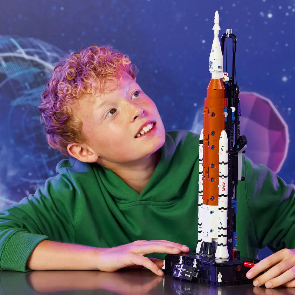 LEGO Technic NASA Artemis Space Launch System lifestyle image shows a child carefully attaching rocket sections, focusing on alignment and details, with a colorful space-themed backdrop behind the build area.