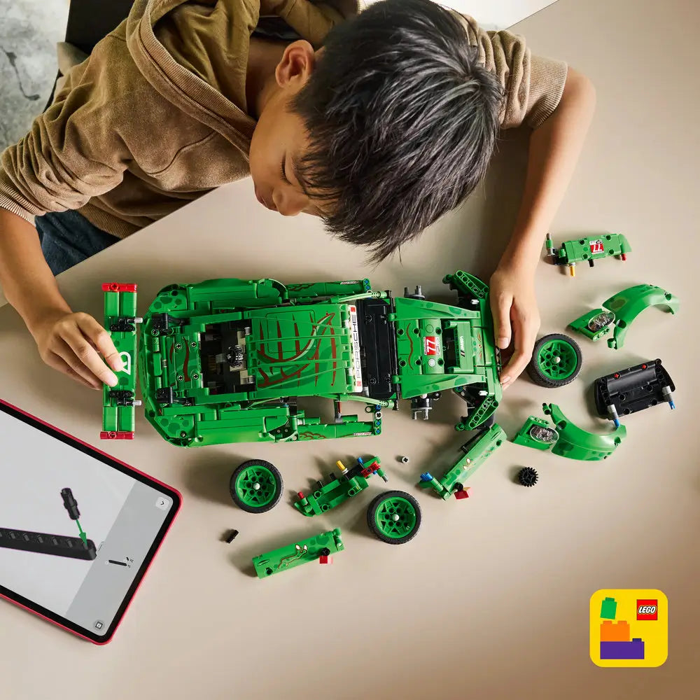 LEGO Technic Porsche 911 GT3 R building scene shows green plastic Technic chassis and loose components spread on a table as a child assembles the race car using a digital instruction app in a bright indoor setting.
