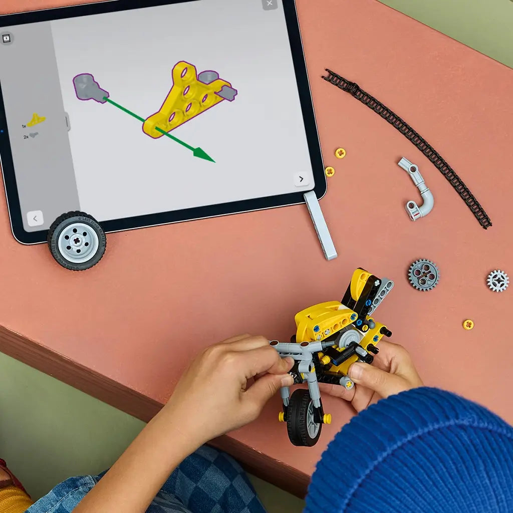 LEGO Technic yellow motorcycle build scene shows yellow and gray plastic components, black chain pieces, and small parts arranged on a table, illustrating assembly process with digital instructions on a tablet in a creative workspace.