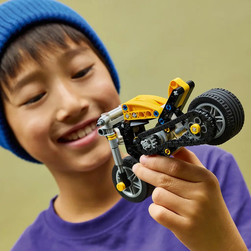 LEGO Technic yellow motorcycle toy shows a bright yellow and gray plastic bike with black frame and rubber tires, held by a smiling child, demonstrating hands-on play and building experience in a casual indoor setting.