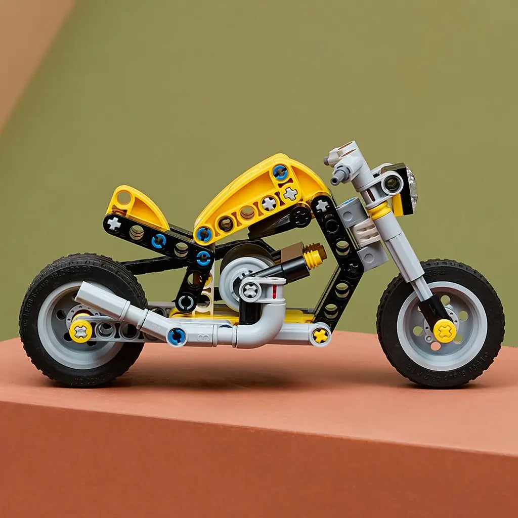 LEGO Technic yellow motorcycle display features a yellow and gray plastic bike with black frame, rubber tires, and visible engine details, positioned on a tabletop in a softly lit indoor environment for showcase.