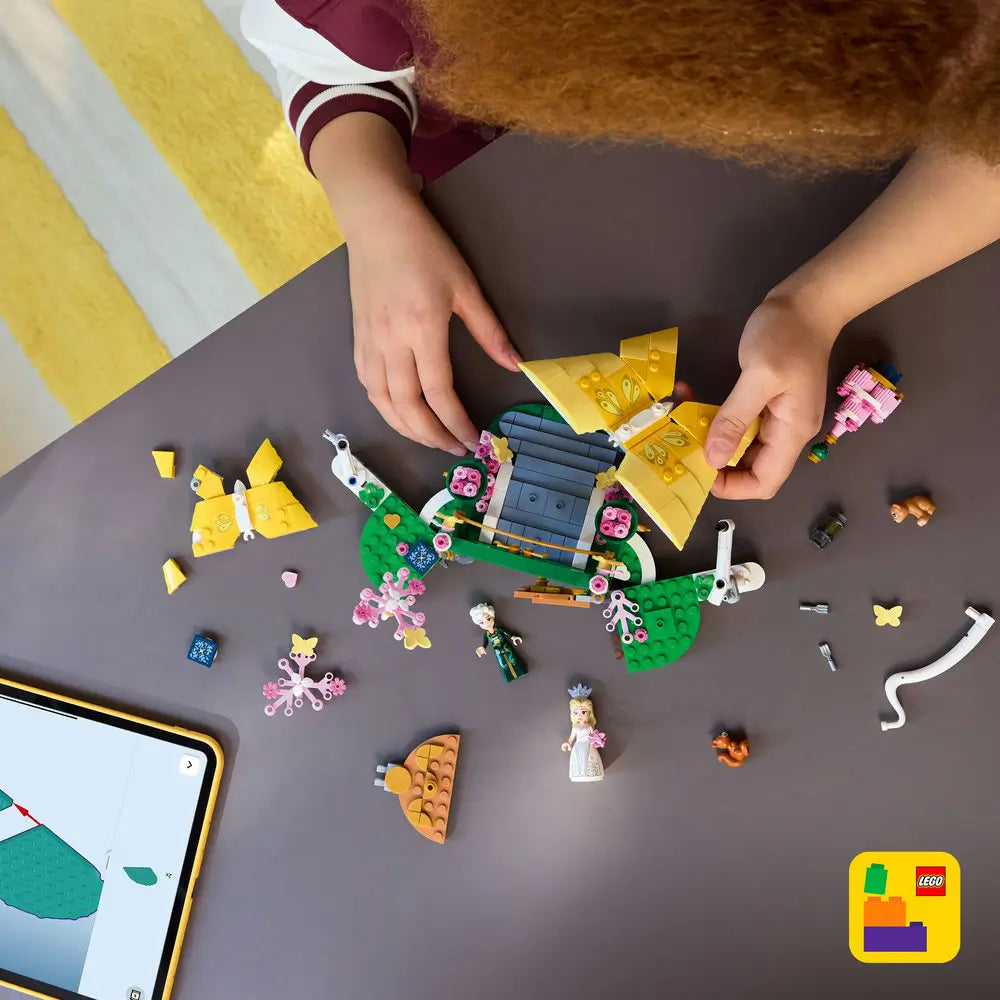 LEGO Wicked Glinda's Wedding Day playset shows a child assembling yellow butterfly models, garden pieces and mini-dolls beside digital building instructions on an indoor tabletop with detailed LEGO elements and realistic display presentation.