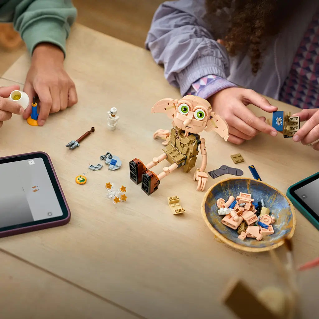 LEGO Harry Potter Dobby the Free Elf build in tan plastic bricks sits partly assembled on a table while children handle accessories beside loose pieces, tablets and a parts bowl.