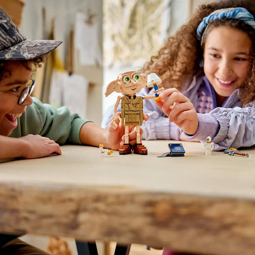 LEGO Harry Potter Dobby the Free Elf figure in tan plastic bricks stands on a table as two children play with a small lamp, blue book, skeleton and sparkling star accessories.
