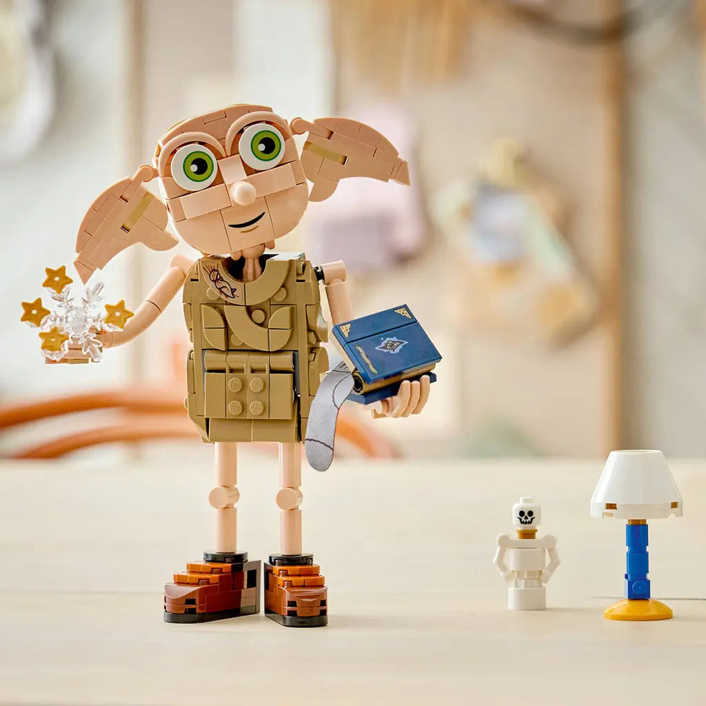 LEGO Harry Potter Dobby the Free Elf display in tan plastic bricks, with large green eyes and brown shoes, holding gold star effects and a blue book beside a lamp and skeleton.