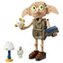 LEGO Harry Potter Dobby the Free Elf figure in tan plastic bricks, with large green eyes and brown shoes, holding gold star effects and a blue book beside a lamp and skeleton.