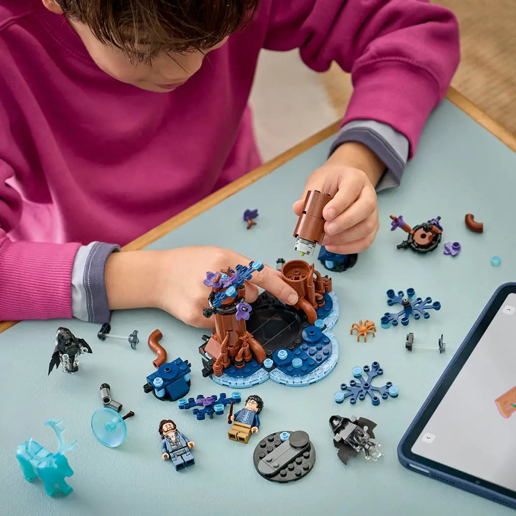 LEGO Harry Potter Forbidden Forest building scene shows a child in a pink top attaching a brown tree trunk amid blue branches, minifigures, loose bricks, and a tablet.