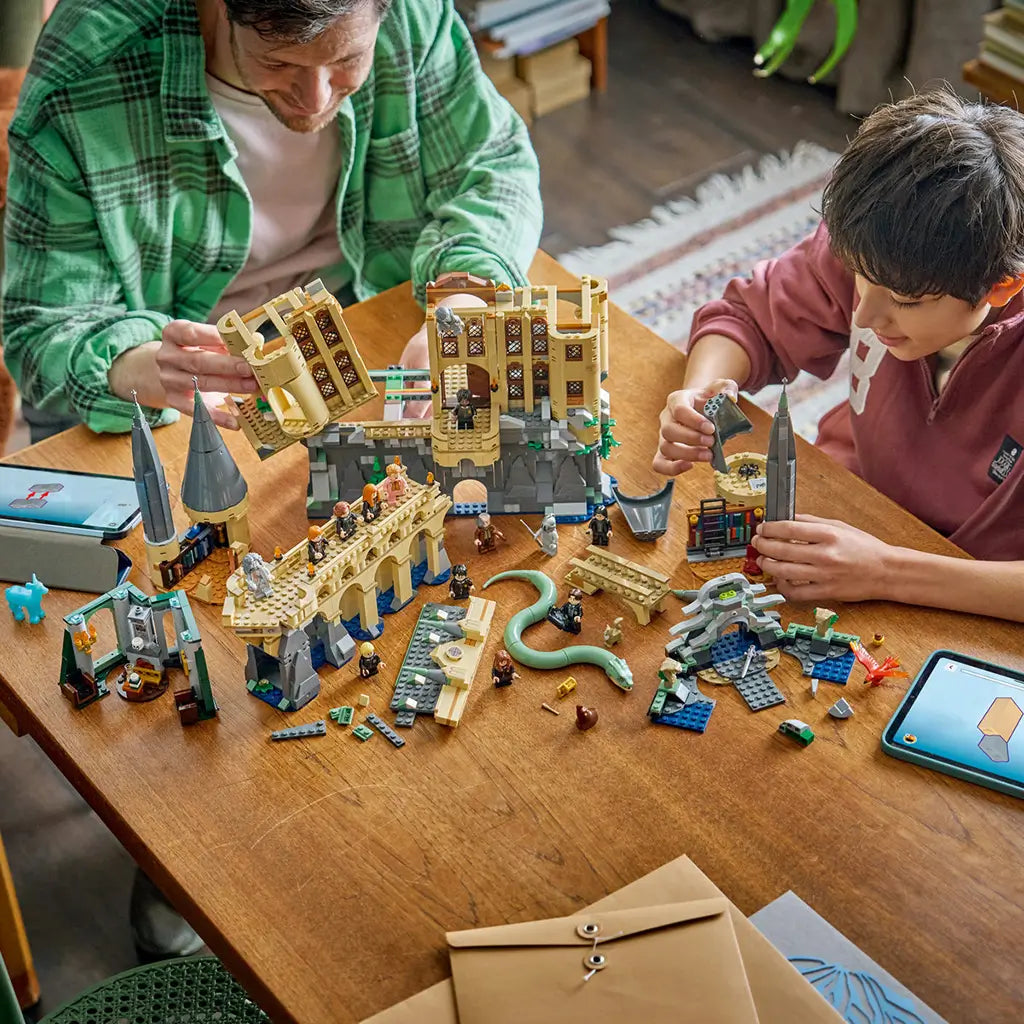 LEGO Harry Potter Hogwarts Castle East Wing building scene shows an adult and child assembling tan and gray castle sections, towers, bridge, minifigures, and basilisk.