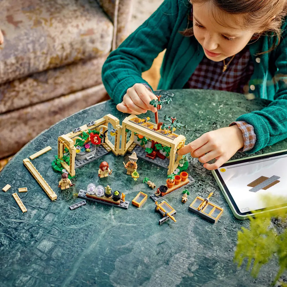 LEGO Harry Potter Herbology Class playset shows a child building the tan greenhouse with magical plants, minifigures and accessories beside a tablet on an indoor table with detailed LEGO elements and realistic display presentation.
