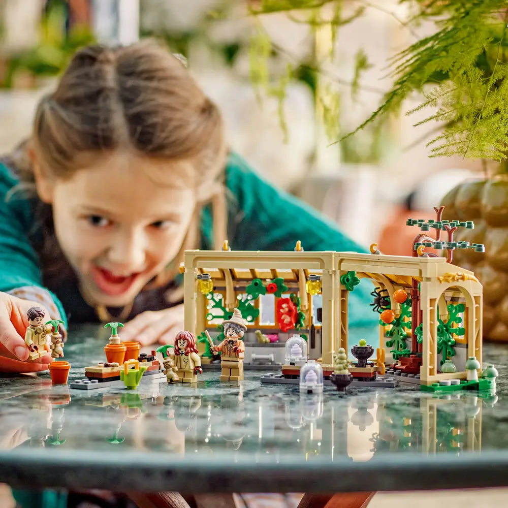 LEGO Harry Potter Herbology Class playset features a child playing with the tan greenhouse, Professor Sprout and student minifigures on a reflective indoor tabletop with plants behind with detailed LEGO elements and realistic display presentation.