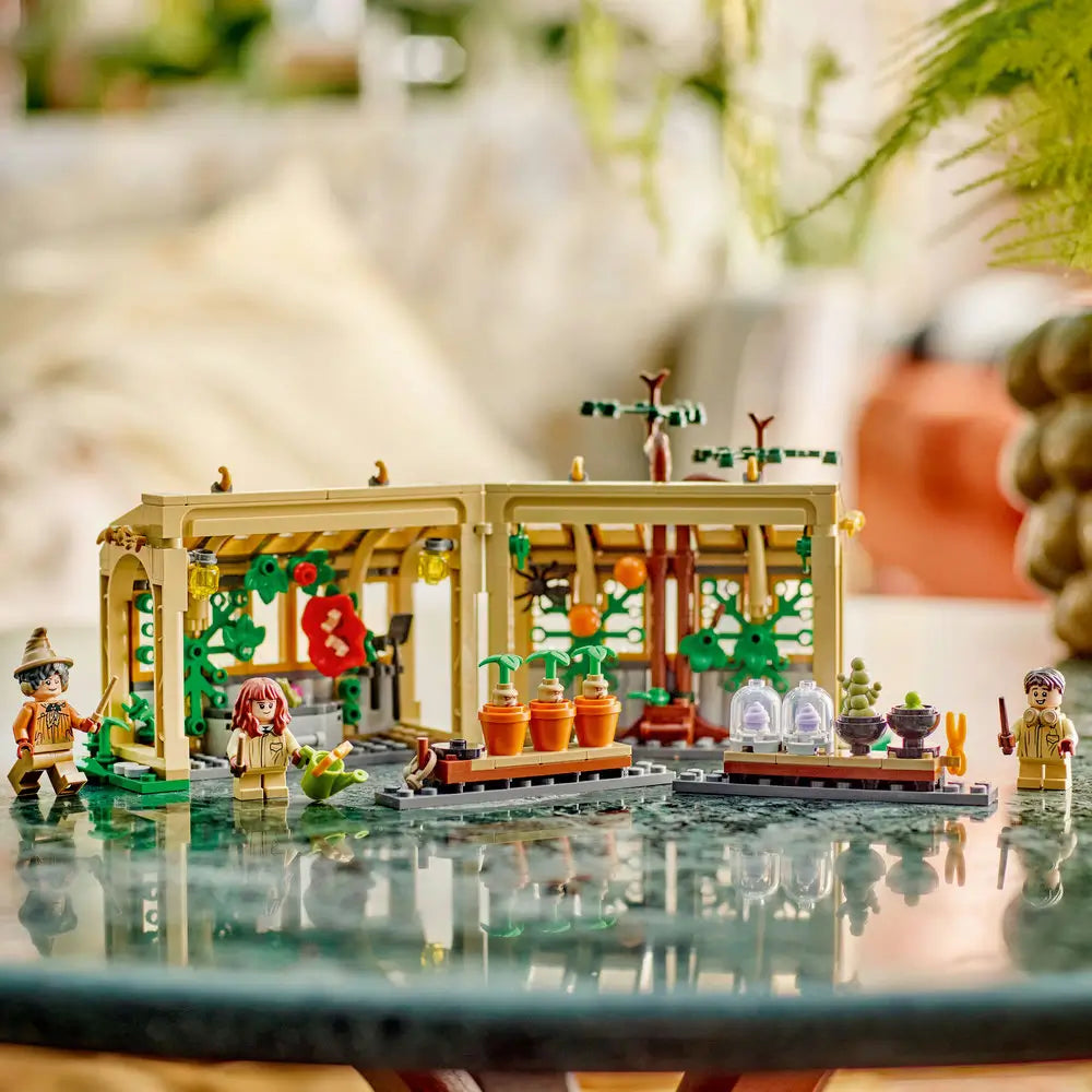 LEGO Harry Potter Herbology Class playset displays the tan greenhouse, magical plants, classroom accessories and minifigures arranged on a reflective indoor tabletop with greenery behind with detailed LEGO elements and realistic display presentation.