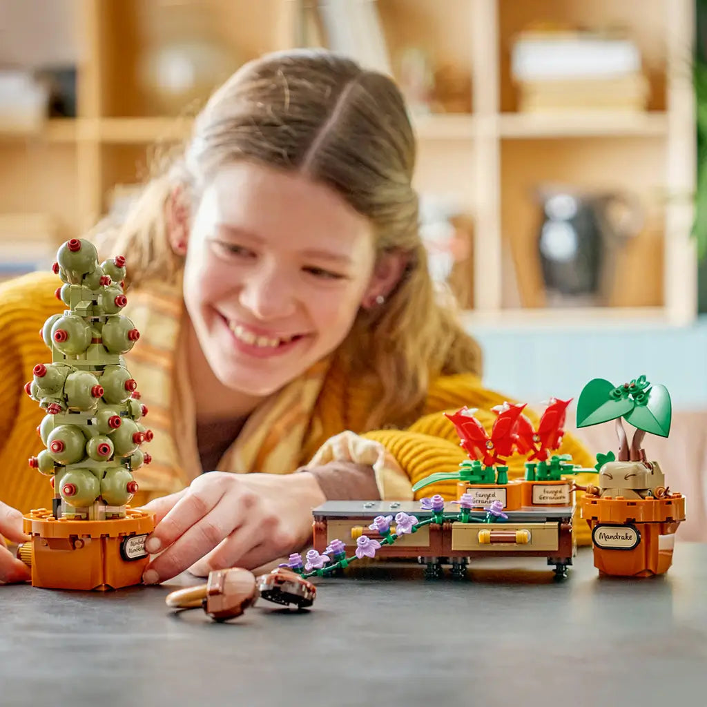 LEGO Harry Potter Hogwarts Herbology Plants play scene shows a smiling child with green and red brick plants in brown pots, including a Mandrake and Mimbulus Mimbletonia.